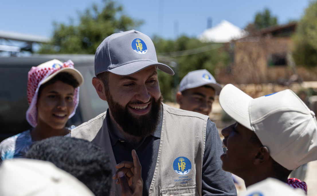 Black-and-white photograph of Sheikh Majed Mahmoud in a grey cap and utility vest, smiling as he greets a child at the Suleiman Center