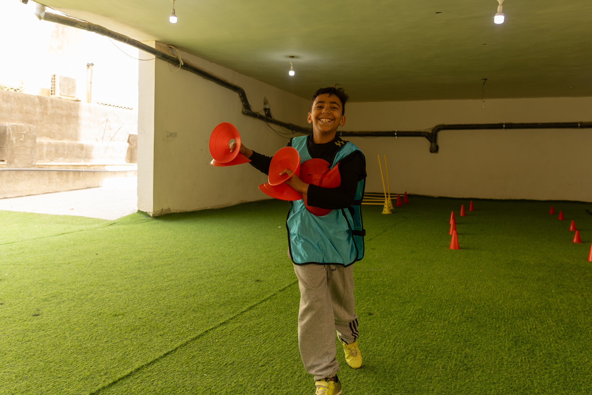 A boy on a turf field holding sports cones, smiling