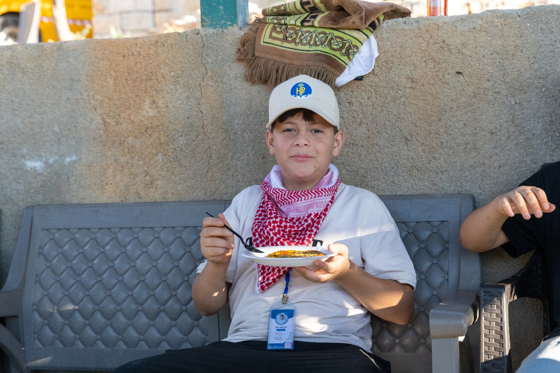 A boy in an HDF cap and keffiyeh eating a meal on a bench